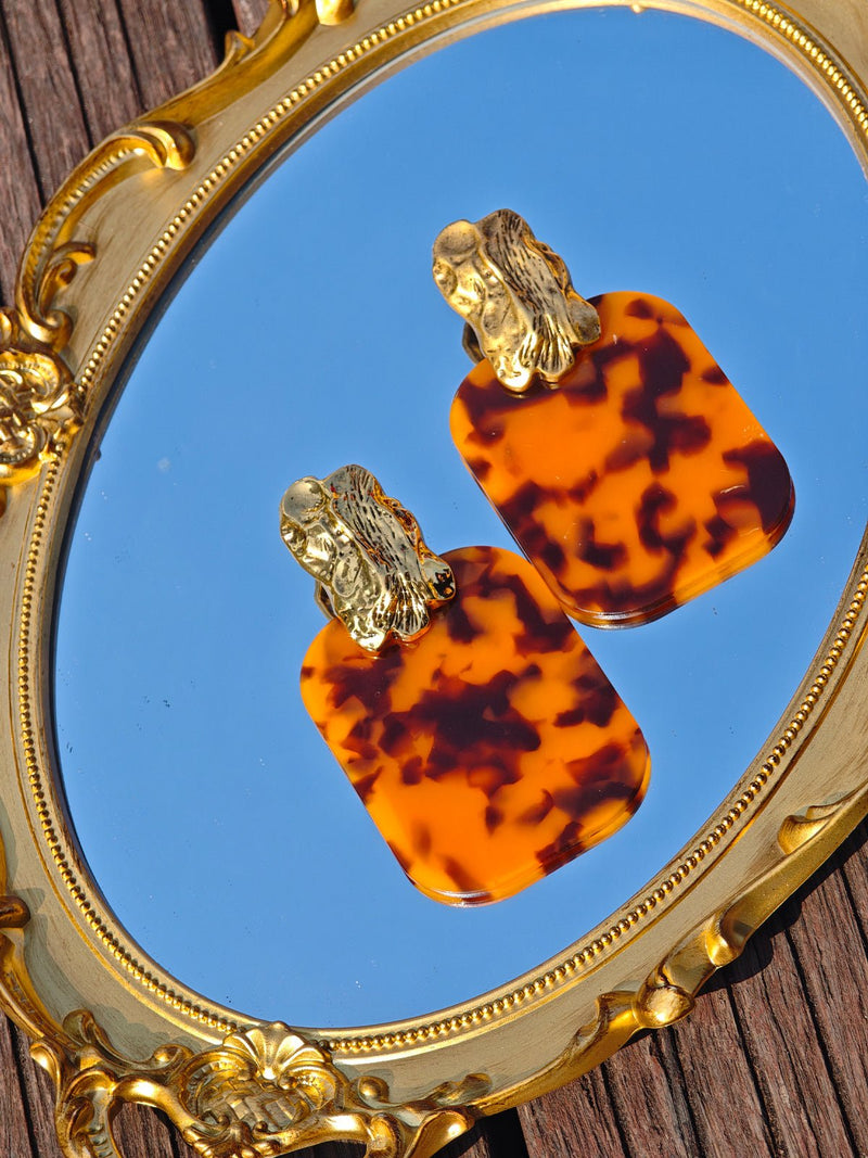 The Social Queen Leopard-Print Acrylic Earrings, featuring gold-plated inlay, are displayed on a gold-framed mirror resting on a wooden surface.