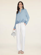 A woman stands against a plain background wearing Social Queen's Linen Straight-Leg Pants in white, paired with a blue lace-trim blouse, silver heels, a small silver handbag, and statement earrings.