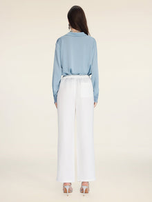 A woman faces away, wearing Social Queen's Linen Straight-Leg Pants paired with a light blue long-sleeve blouse and white heeled sandals, set against a plain background.