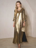 A woman stands indoors in the Social Queen Metallic Crystal-Embellished Slit Dress, paired with a gold handbag, matching gold earrings, and sandals.