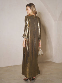 A woman stands indoors facing away, wearing the Social Queen Metallic Crystal-Embellished Slit Dress with three-quarter sleeves and an elegant silhouette. She holds a small handbag.