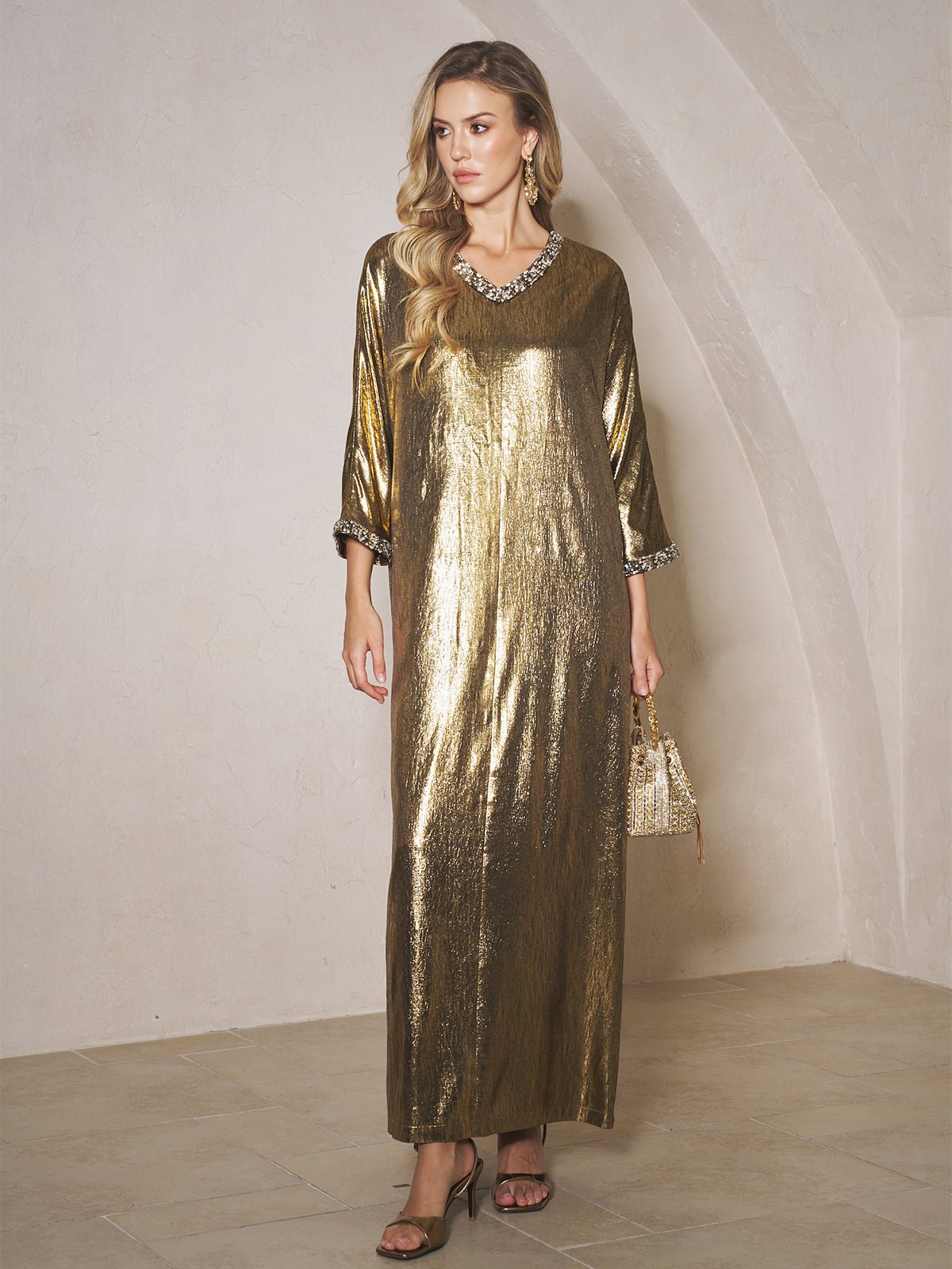 A woman models the Social Queen Metallic Crystal-Embellished Slit Dress with three-quarter sleeves, holding a gold handbag and wearing heeled sandals indoors.