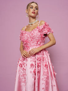 A woman models the Social Queen Off-Shoulder Floral Satin Gown, featuring delicate 3D floral details, standing against a pink backdrop with drop earrings and a choker, gazing slightly to the side.