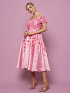 A woman stands against a pink background wearing the Social Queen Off-Shoulder Floral Satin Gown with 3D floral embellishments, paired with strappy heels.