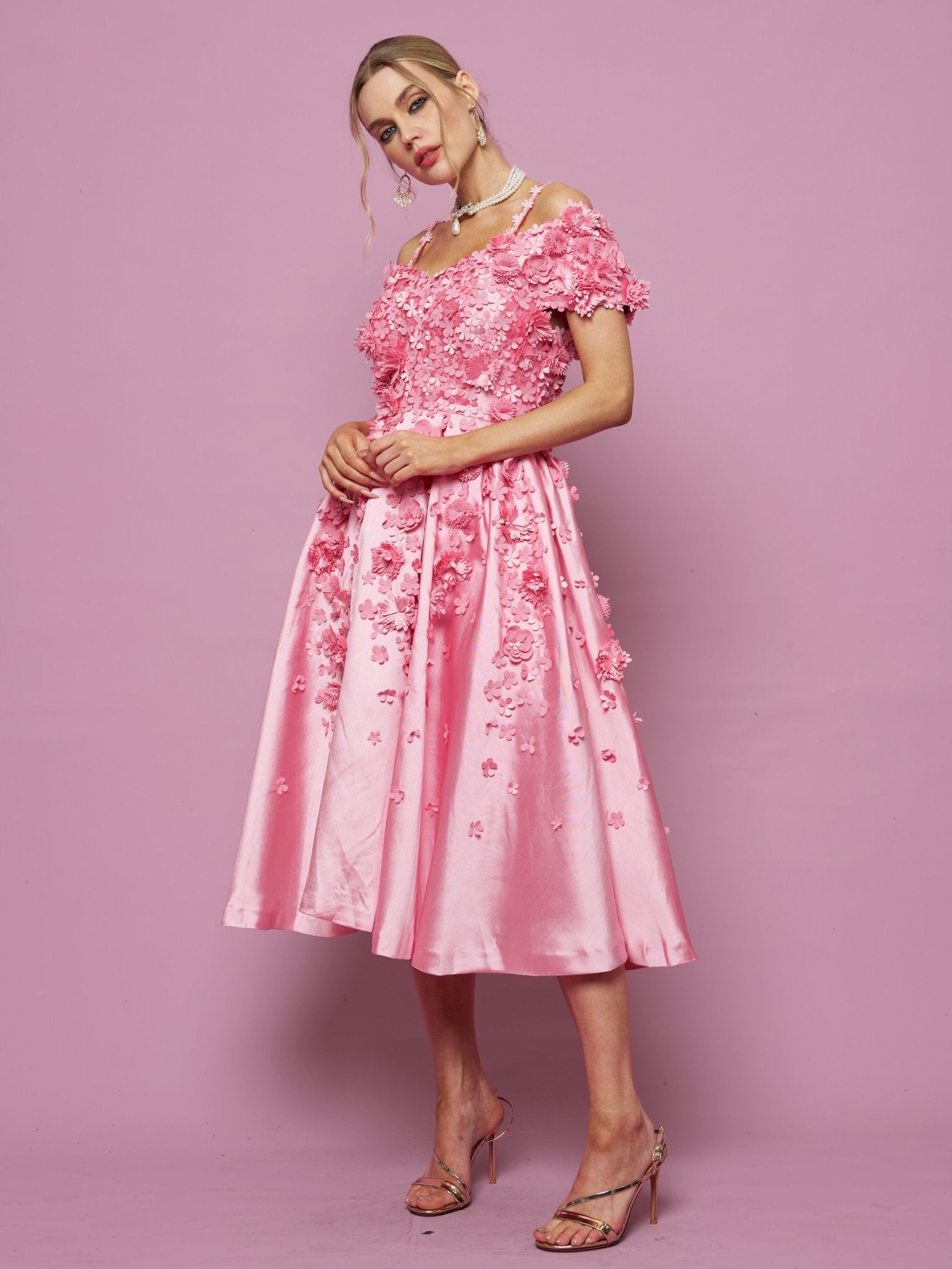 A woman stands against a pink background wearing the Social Queen Off-Shoulder Floral Satin Gown with 3D floral embellishments, paired with strappy heels.