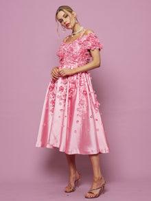 A woman stands against a pink background wearing the Social Queen Off-Shoulder Floral Satin Gown with 3D floral embellishments, paired with strappy heels.