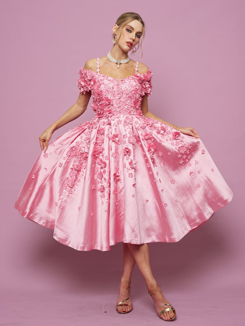A woman poses against a pink background in the Social Queen Off-Shoulder Floral Satin Gown, featuring 3D floral details, paired with silver sandals.