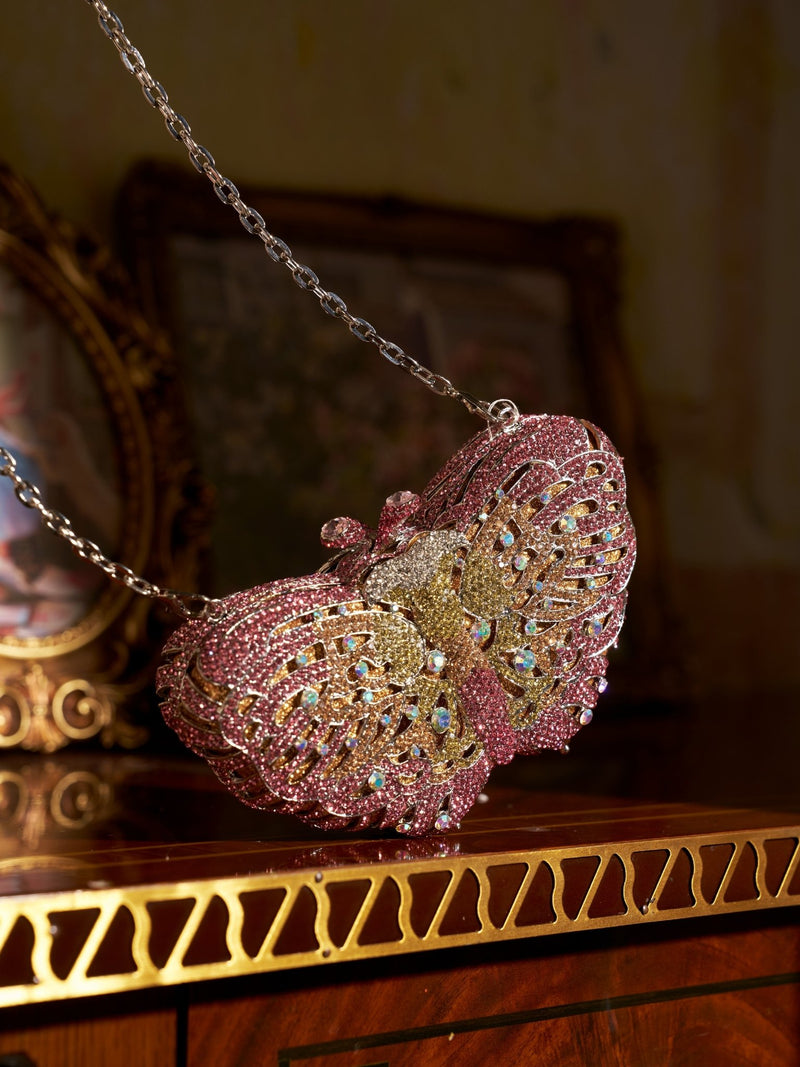 The Social Queen Ombré Crystal Butterfly Mini Bag, featuring a chain strap, rests on polished wood with gold detailing in front of a framed picture—a dazzling evening clutch ideal as a wedding accessory.