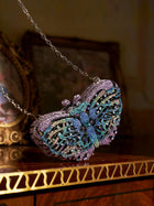The Social Queen Ombré Crystal Butterfly Mini Bag, adorned with blue, purple, and green crystals on a chain strap, sits atop a polished wooden surface—the ideal wedding or evening accessory.