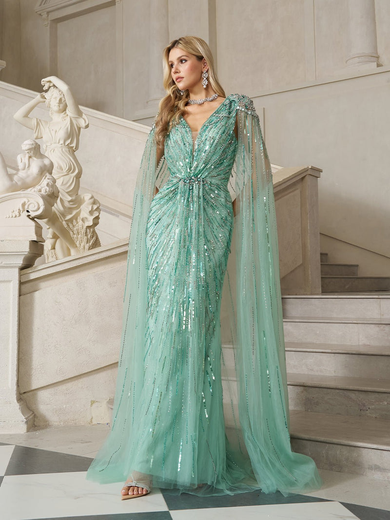 A woman in Social Queen's Ombré Sequin Cape Gown with Beaded Fringe stands on a marble staircase beside a classical statue, the gown’s shimmering fabric and elegant cape sleeves catching the light.