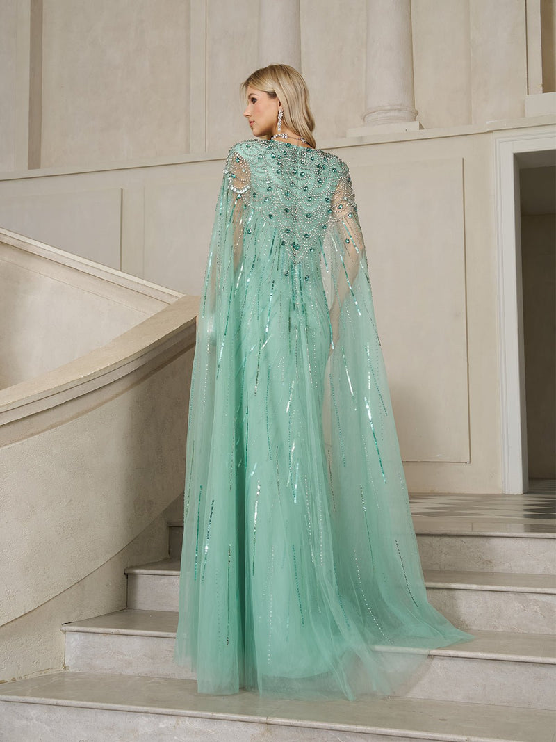 A woman in the Social Queen Ombré Sequin Cape Gown with Beaded Fringe stands on a staircase, facing away—the gown’s sheer cape sleeves and shimmering beaded details create an ethereal, elegant look.