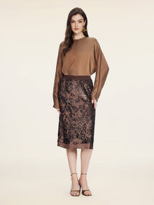 A woman faces forward against a plain background, wearing a long-sleeved brown blouse, gold earrings, brown heeled sandals, and the Social Queen Ombre Sequin Velvet Skirt.