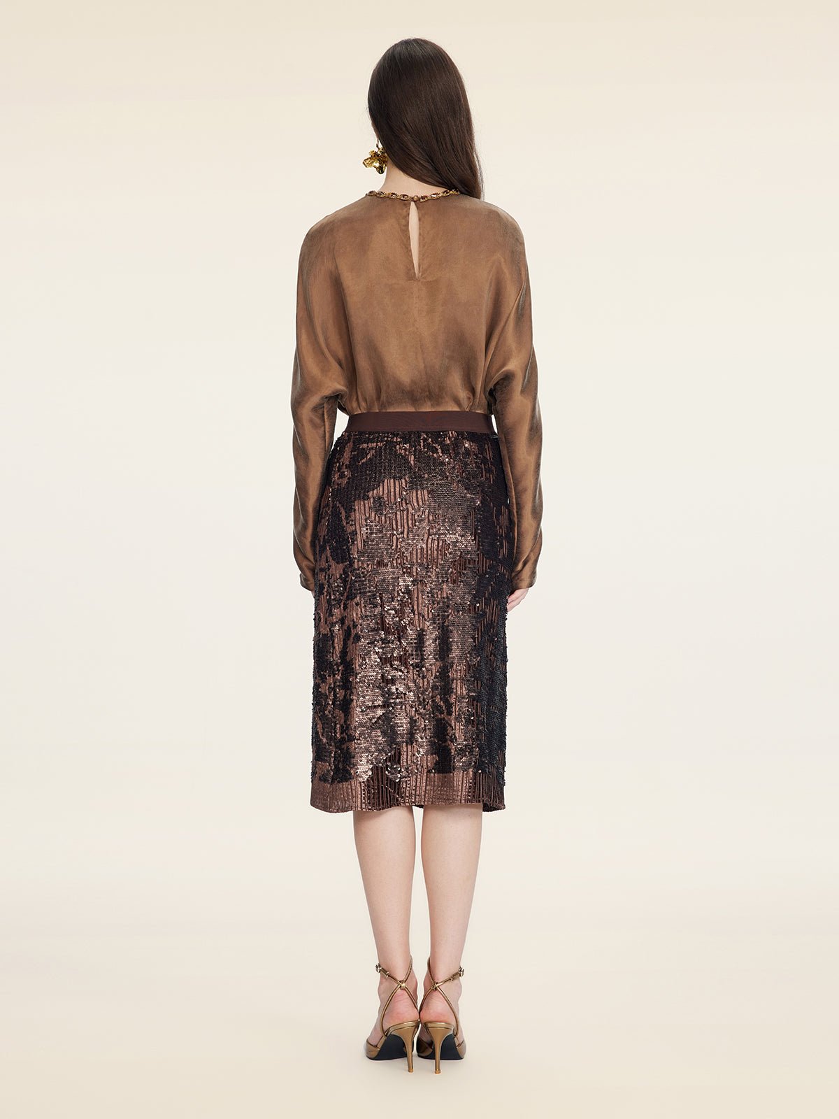 A woman stands facing away, wearing a brown long-sleeve blouse, Social Queen’s Ombre Sequin Velvet Skirt with hand-shaped pleats, and strappy high-heeled sandals against a plain background.