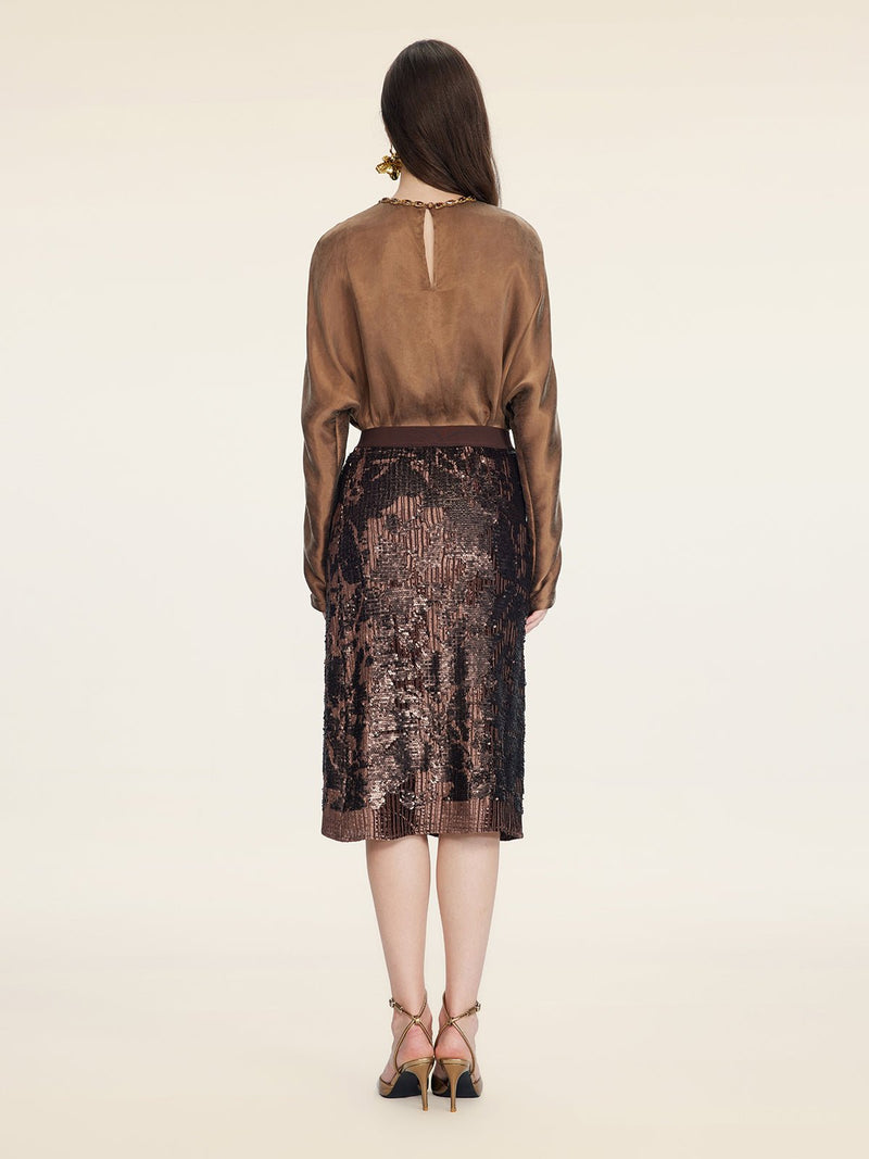 A woman stands facing away, wearing a brown long-sleeve blouse, Social Queen’s Ombre Sequin Velvet Skirt with hand-shaped pleats, and strappy high-heeled sandals against a plain background.