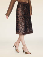 A woman poses against a neutral background wearing a brown long-sleeve blouse, gold high-heeled sandals, and the Social Queen Ombre Sequin Velvet Skirt.