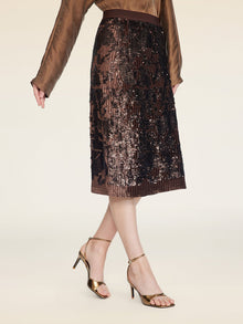 A woman poses against a neutral background wearing a brown long-sleeve blouse, gold high-heeled sandals, and the Social Queen Ombre Sequin Velvet Skirt.