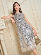 A woman in the Social Queen Ombre Silver & Gold Sequin Embellished Dress with a diamond-trimmed neckline stands indoors, leaning on a mantel and holding a small gold handbag.