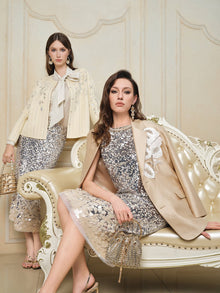 Two women showcase the Social Queen Ombre Silver & Gold Sequin Embellished Dress, featuring sparkling details and a diamond-trimmed neckline, as they pose indoors by a cream ornate sofa against a beige wall.