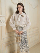 A woman stands indoors in a shimmery blouse with a bow at the neck, wearing the Ombre Silver & Gold Sequin Slim Skirt by Social Queen, and holding a small gold purse against an ornate cream-colored wall.