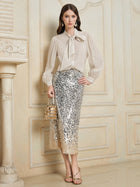 A woman stands indoors wearing a sheer white blouse with a bow and the Social Queen Ombre Silver & Gold Sequin Slim Skirt, holding a gold handbag and wearing gold sandals.