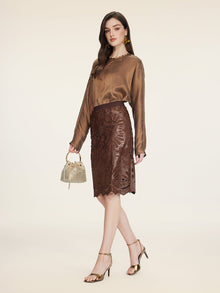 A woman wears Social Queen’s Openwork Embroidered Leather Skirt—a knee-length piece with a wavy hem—paired with a brown satin blouse, gold heels, and a small gold handbag against a plain background.