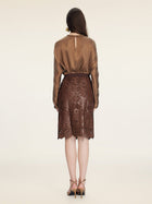 A woman faces away in a sheer brown blouse and the Social Queen Openwork Embroidered Leather Skirt with hand-embroidered details and a wavy hem, paired with heeled sandals, set against an off-white background.