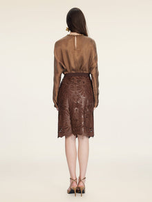 A woman faces away in a sheer brown blouse and the Social Queen Openwork Embroidered Leather Skirt with hand-embroidered details and a wavy hem, paired with heeled sandals, set against an off-white background.