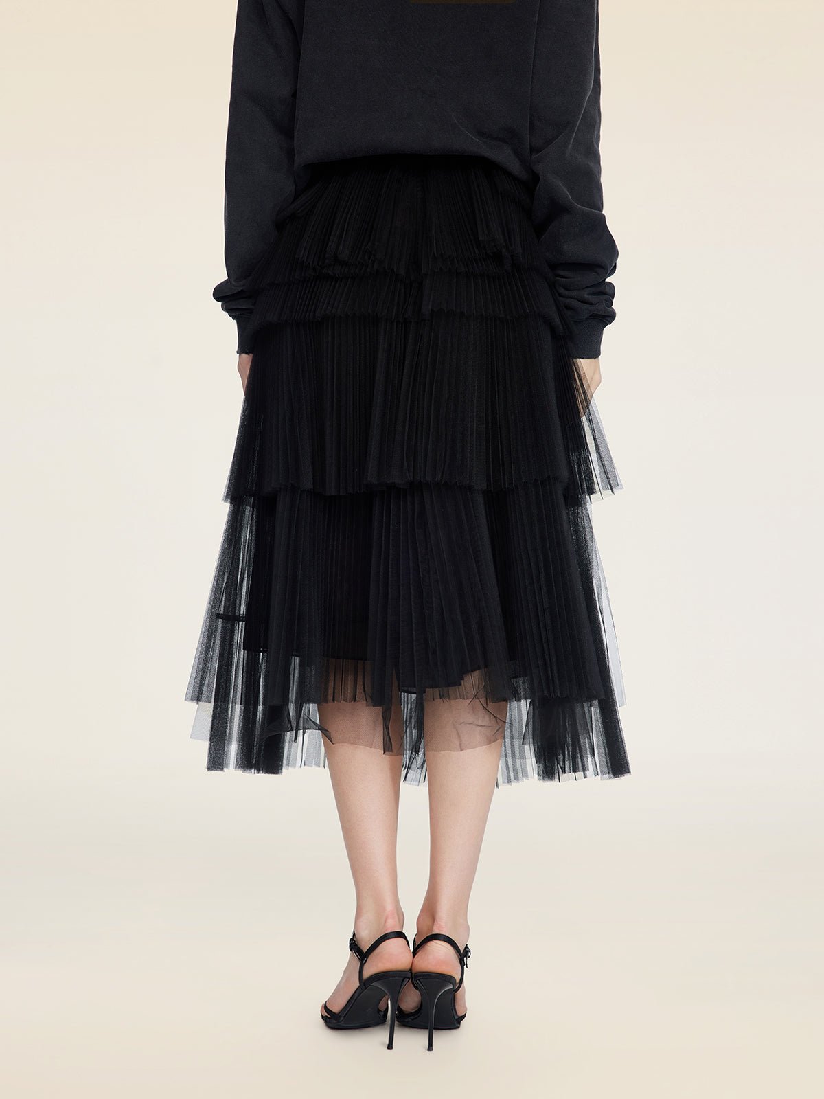 A person stands facing away against a plain light background, wearing the Social Queen Organza Pleated Skirt, a black long-sleeve top, and black heeled sandals.