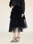 A person stands against a plain light background wearing the Social Queen Organza Pleated Skirt with layered pleats, a black long-sleeve top, and black strappy heels.