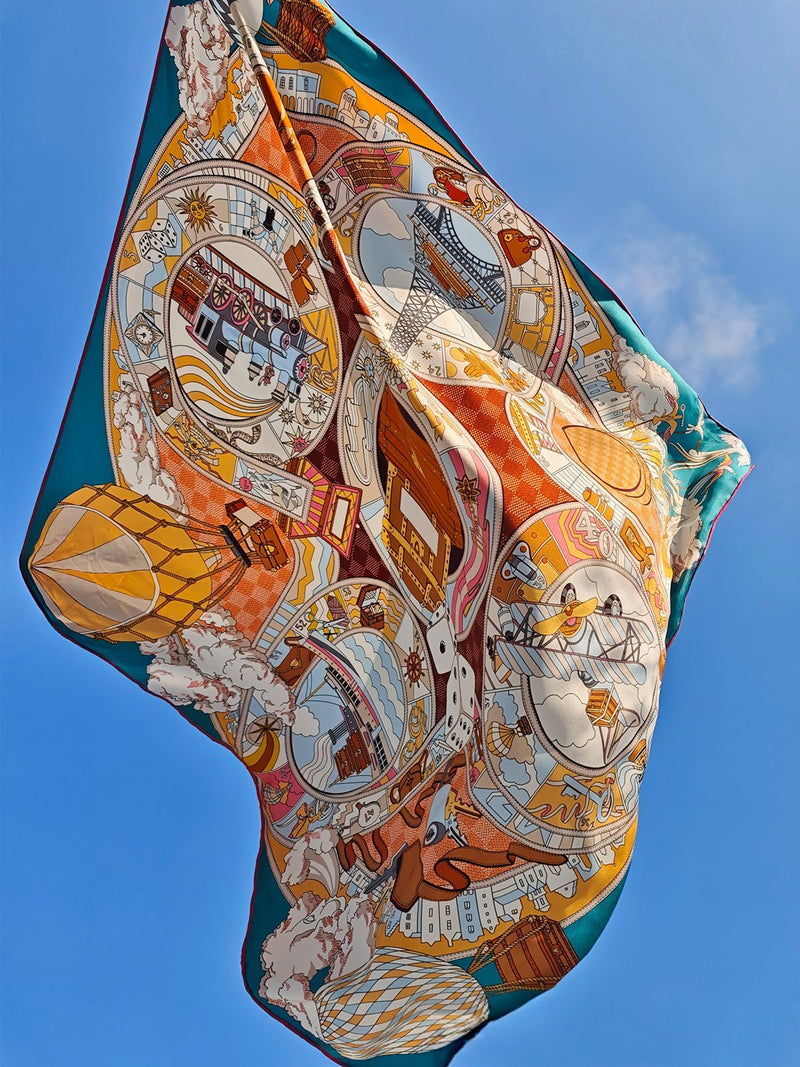 The Social Queen Original Pattern Print Silk Scarf, featuring buildings, hot air balloons, and travel motifs, billows against a clear blue sky.