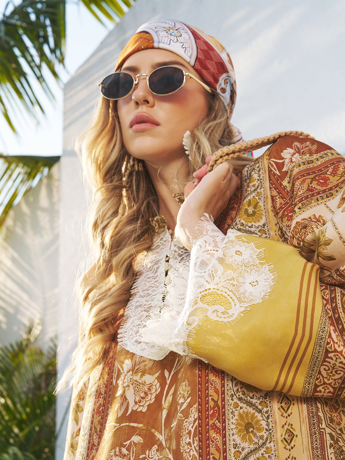 A woman stands outdoors by a sunlit white wall with palm leaves, wearing round sunglasses and the Social Queen Original Pattern Print Silk Scarf with a floral, lace-trimmed outfit.