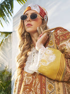 A woman stands outdoors by a sunlit white wall with palm leaves, wearing round sunglasses and the Social Queen Original Pattern Print Silk Scarf with a floral, lace-trimmed outfit.