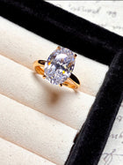 The Social Queen Oval Crystal Ring features a large oval-cut clear crystal set in a gold-tone band, elegantly presented in a beige and black ring box.