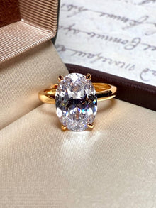 The Social Queen Oval Crystal Ring, featuring a gold-tone adjustable band and an oval-cut clear crystal, is showcased in an open ring box with handwritten paper in the background.