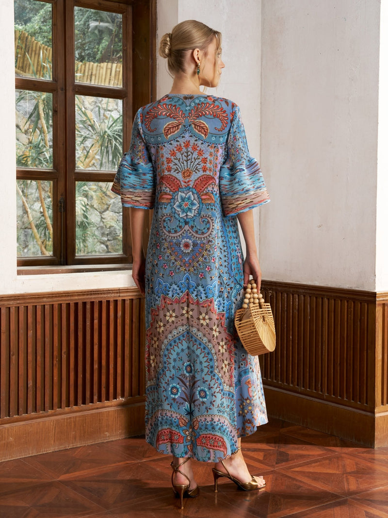A woman indoors faces away, wearing the Social Queen Paisley Print Pleated Midi Dress with flared sleeves and gold heels, holding a woven handbag—an effortlessly chic vacation outfit.