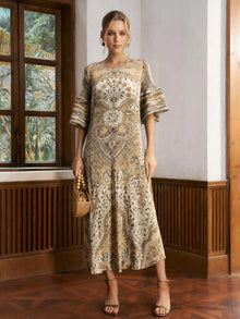 A woman stands indoors wearing the Social Queen Paisley Print Pleated Midi Dress with flared sleeves, holding a woven handbag and pairing it with strappy sandals.