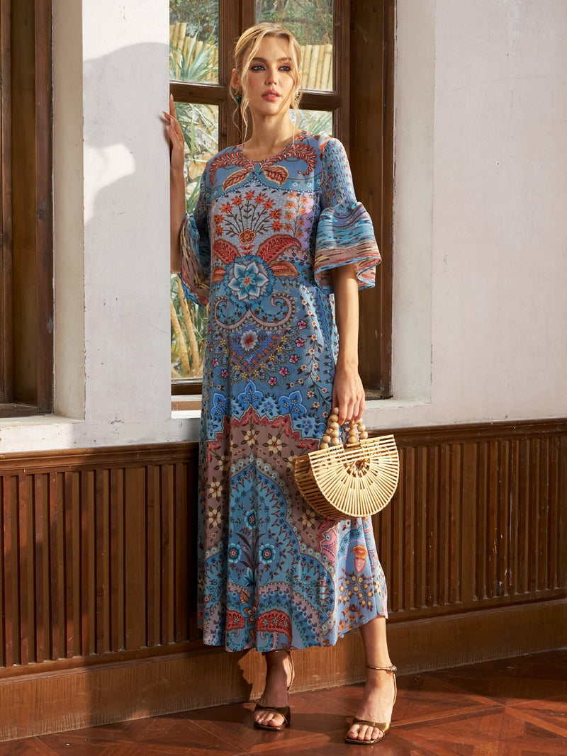A woman stands by a window in the Social Queen Paisley Print Pleated Midi Dress, paired with a wooden handbag and strappy high heels—an effortless vacation-ready look.