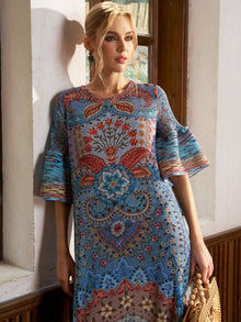 A woman stands by a window in the Social Queen Paisley Print Pleated Midi Dress, holding a round woven bag—an ideal breezy vacation look.