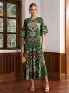 A woman stands indoors wearing the Social Queen Paisley Print Pleated Midi Dress, paired with a woven bag and heeled sandals.
