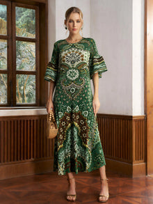 A woman stands indoors wearing the Social Queen Paisley Print Pleated Midi Dress, paired with a woven bag and heeled sandals.