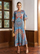 A woman stands indoors wearing the Social Queen Paisley Print Pleated Midi Dress with flared sleeves, paired with a woven handbag and heeled sandals.