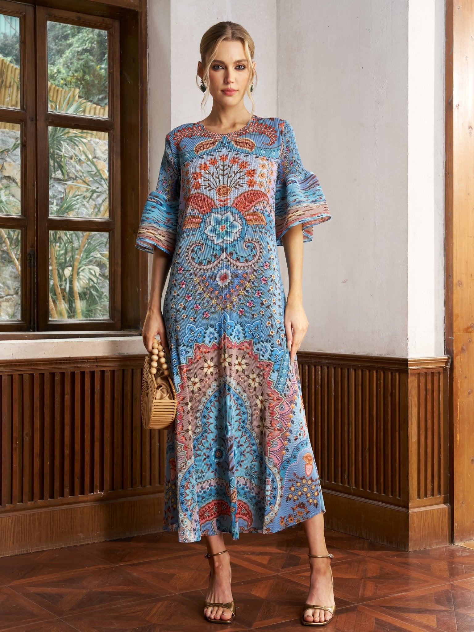 A woman stands indoors wearing the Social Queen Paisley Print Pleated Midi Dress with flared sleeves, paired with a woven handbag and heeled sandals.