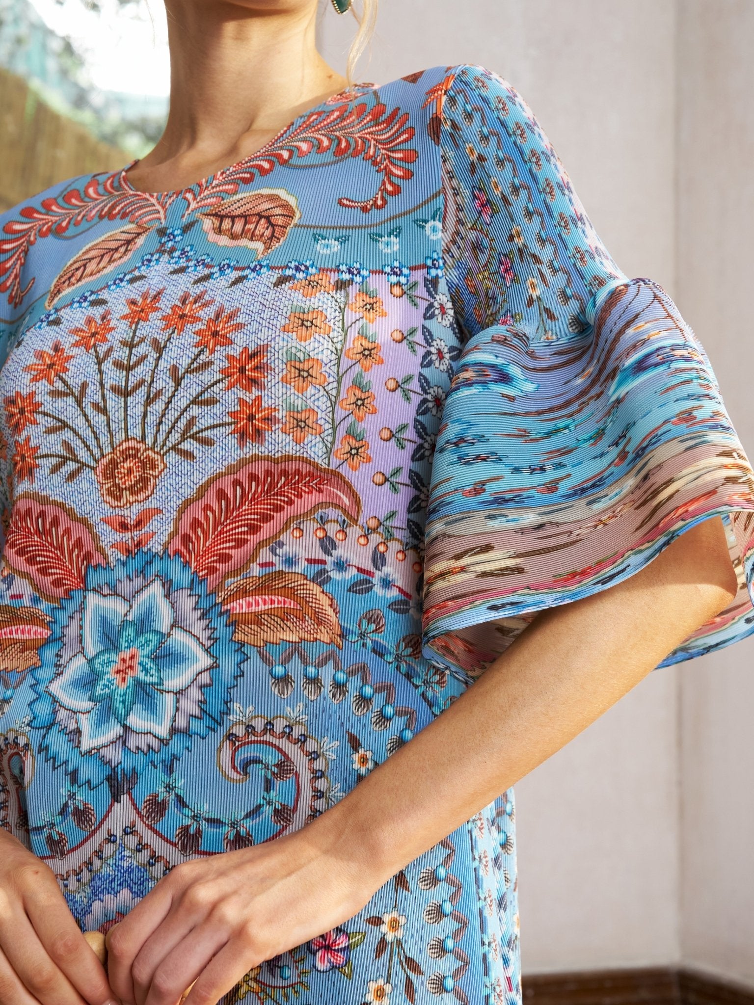 A woman stands indoors near a window wearing the Social Queen Paisley Print Pleated Midi Dress, featuring blue paisley patterns with colorful floral and geometric designs and flared sleeves.