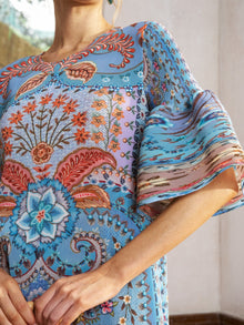 A woman stands indoors near a window wearing the Social Queen Paisley Print Pleated Midi Dress, featuring blue paisley patterns with colorful floral and geometric designs and flared sleeves.