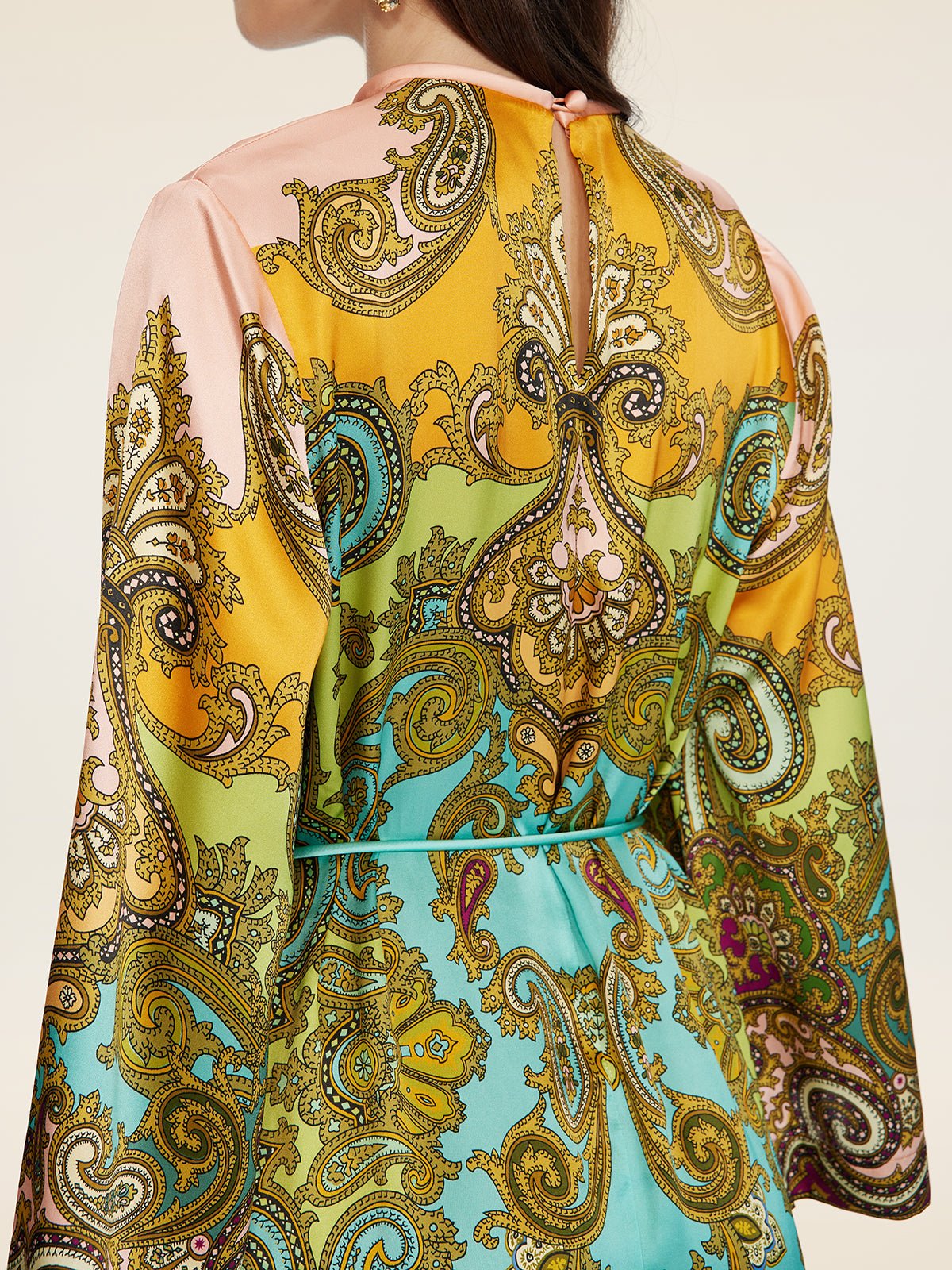 A woman is seen from the back wearing the Social Queen Paisley Printed Silk Satin Dress featuring long sleeves and a vibrant yellow, green, blue, and pink paisley pattern inspired by vintage styles.