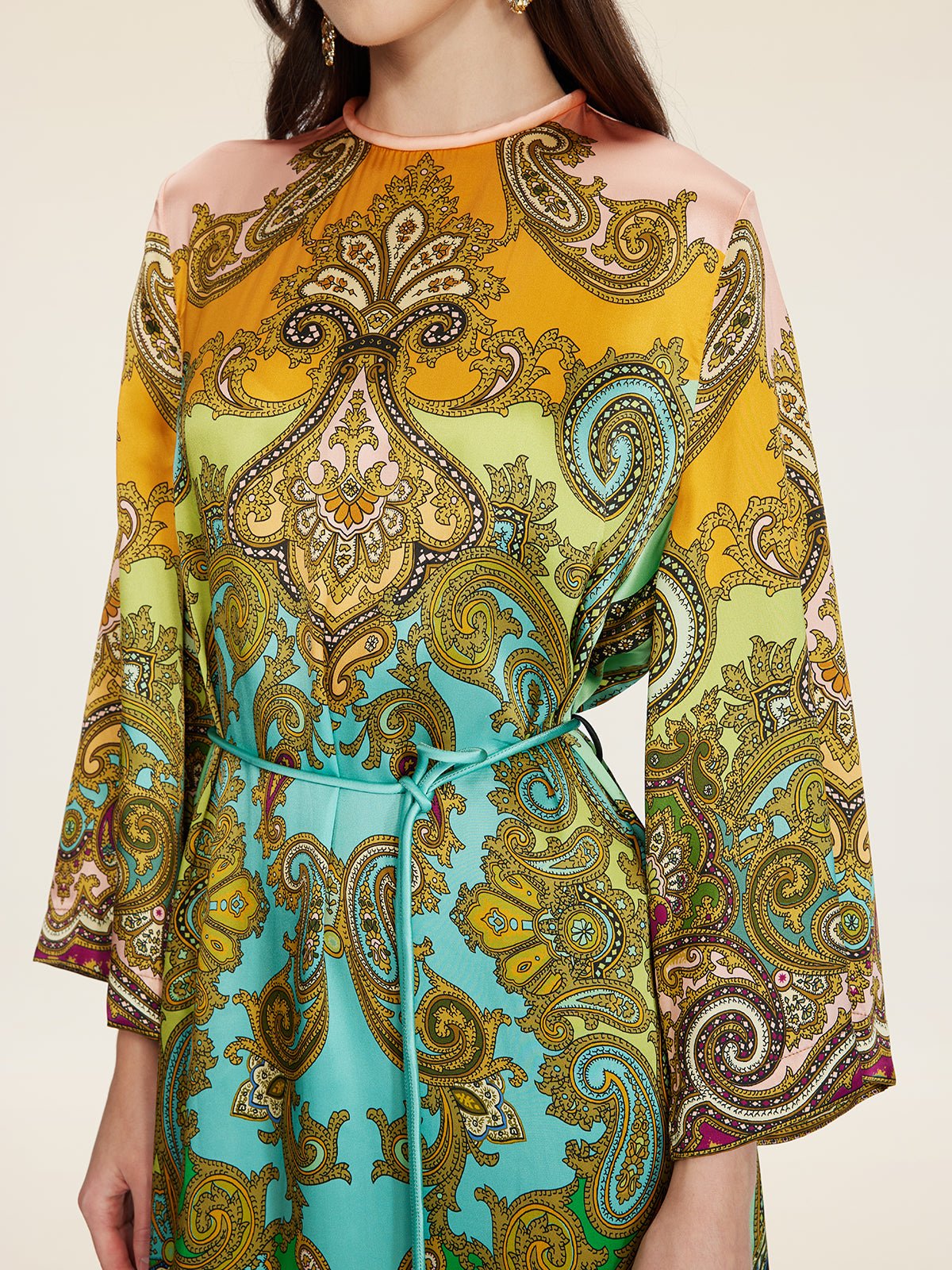 A person wearing the Social Queen Paisley Printed Silk Satin Dress, a vintage-inspired long-sleeved piece with hand-printed yellow, green, and turquoise paisley patterns, cinched at the waist with a thin belt.