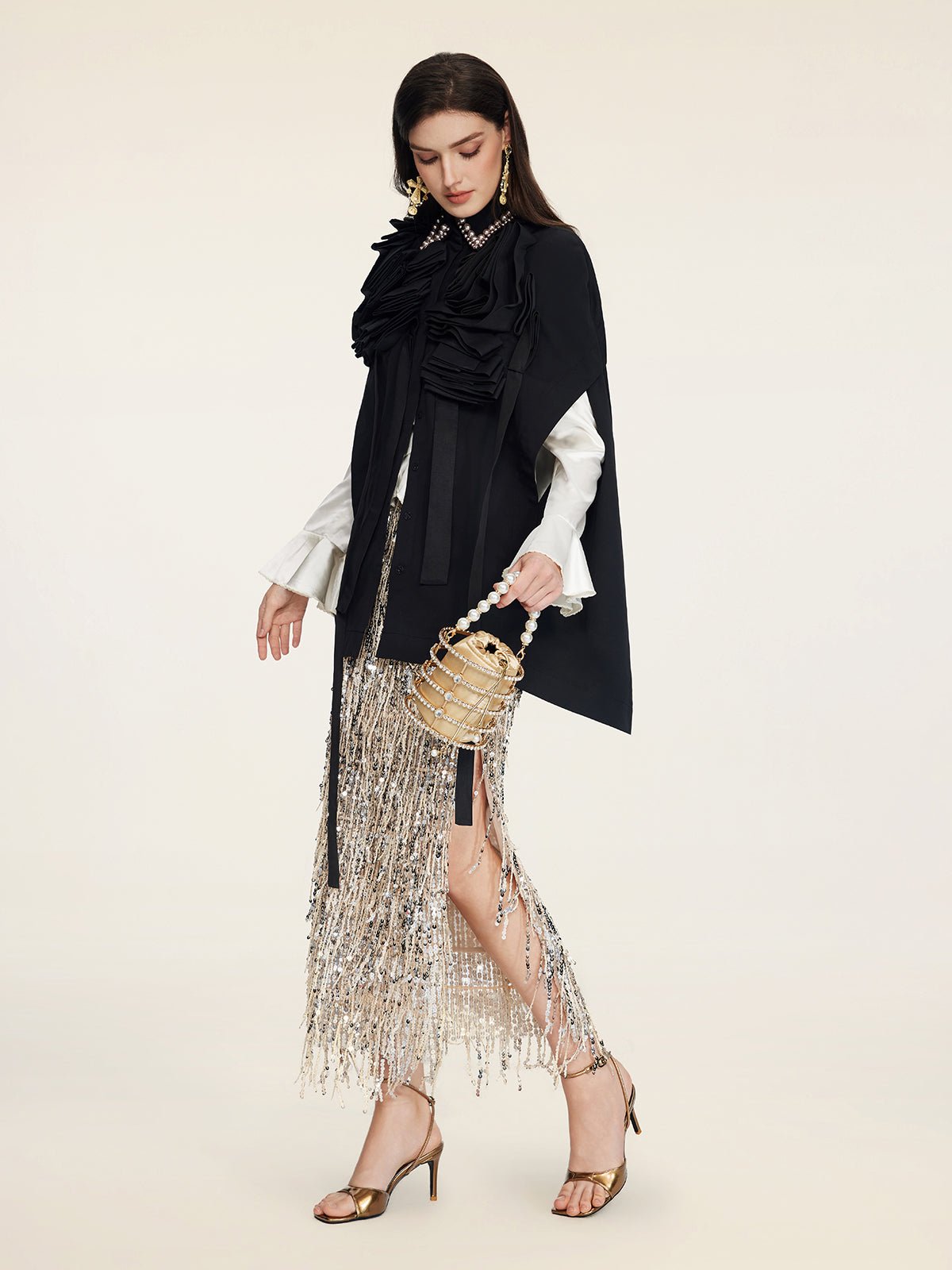 A woman models the Social Queen Pearl & 3D Floral Couture Cape, styled with a white blouse, silver fringe skirt, gold heeled sandals, and a gold handbag against a plain background, showcasing couture craftsmanship.