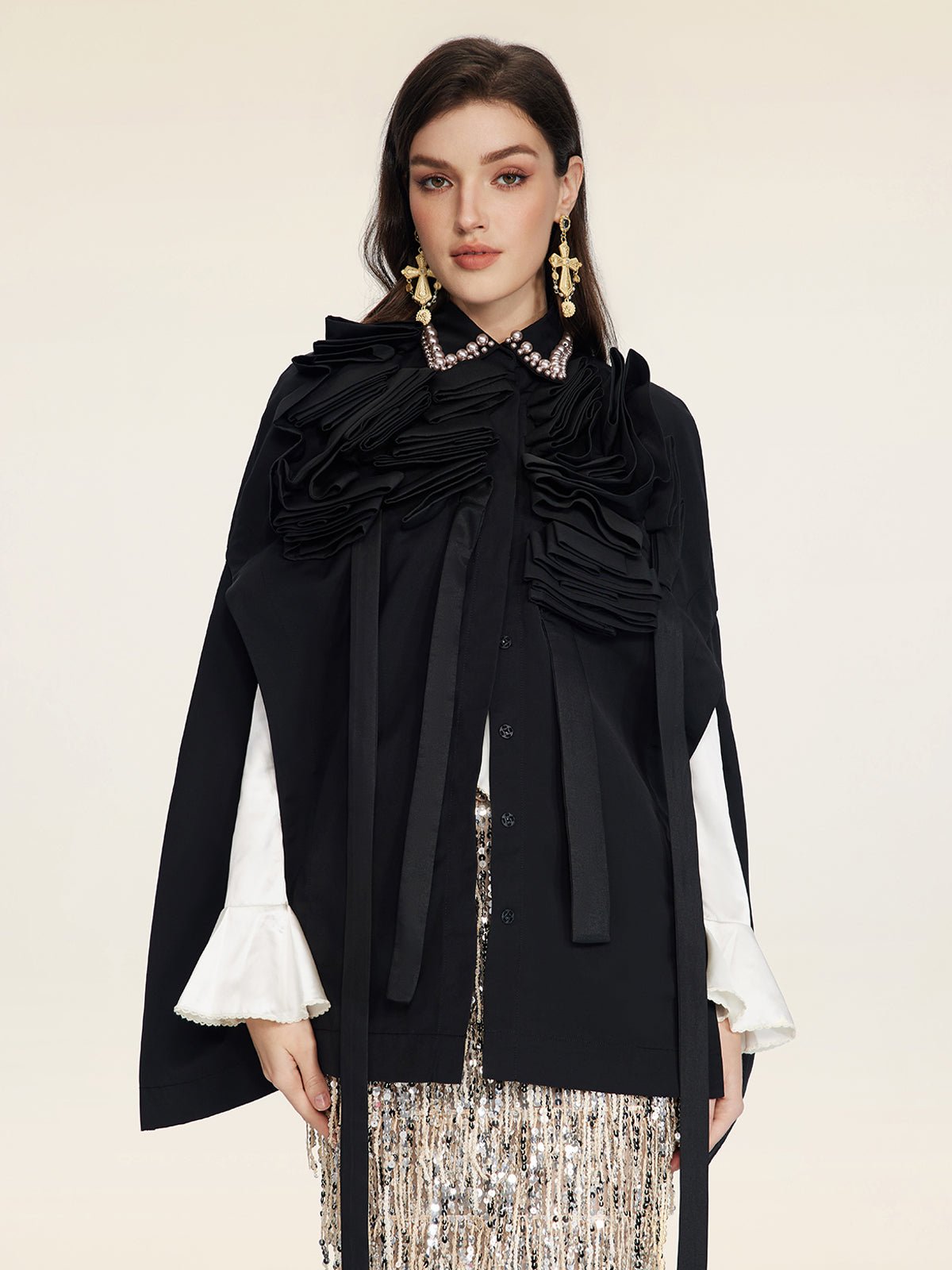 A woman wears the Social Queen Pearl & 3D Floral Couture Cape—black with pearl collar, 3D floral ruffles, and white flared sleeves—styled with statement earrings and a sequined skirt against a plain light background.