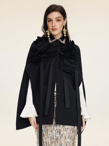 A woman wears the Social Queen Pearl & 3D Floral Couture Cape—black with pearl collar, 3D floral ruffles, and white flared sleeves—styled with statement earrings and a sequined skirt against a plain light background.