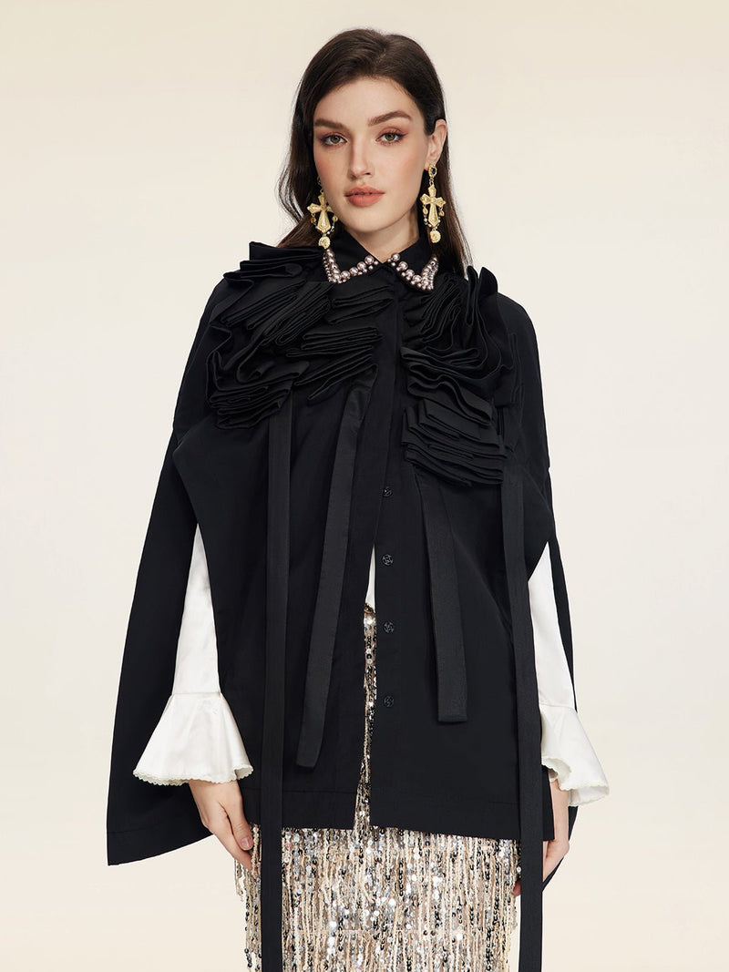 A woman wears the Social Queen Pearl & 3D Floral Couture Cape—black with pearl collar, 3D floral ruffles, and white flared sleeves—styled with statement earrings and a sequined skirt against a plain light background.
